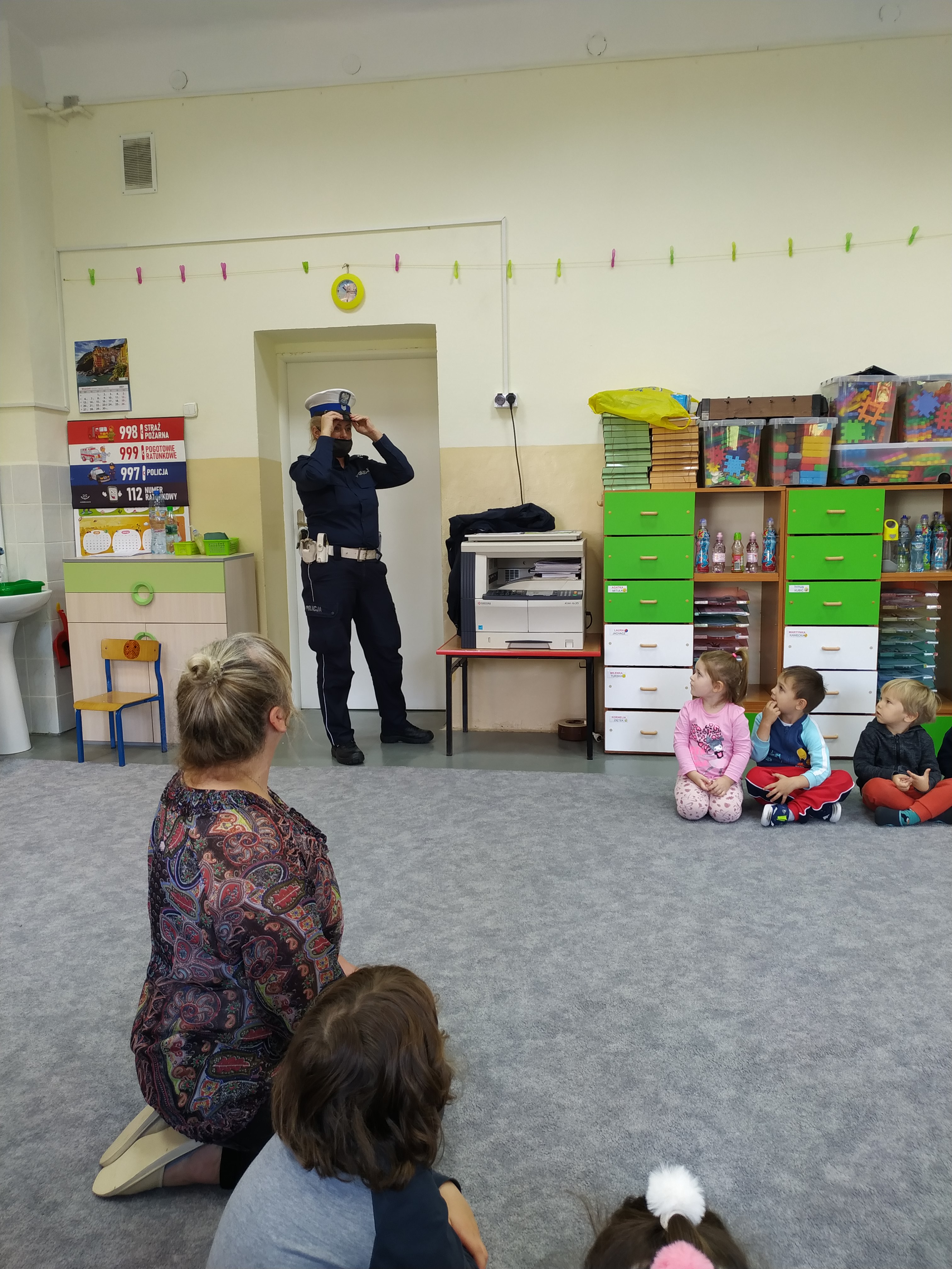 . Dzieci wraz z nauczycielką siedzą w półkolu na dywanie. Przy drzwiach stoi pani policjantka prezantując mundur policyjny.