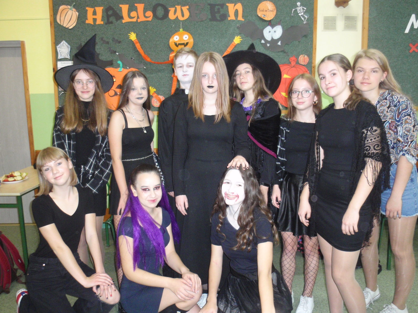 W tle widać gazetkę z okazji Halloween, stoi grupa dziewczynek z jednym chłopcem .Wszyscy ubrani w czarne stroje, 3 dziewczynki mają makijaż na twarzach, Jedna przyczepione fioletowe włosy.