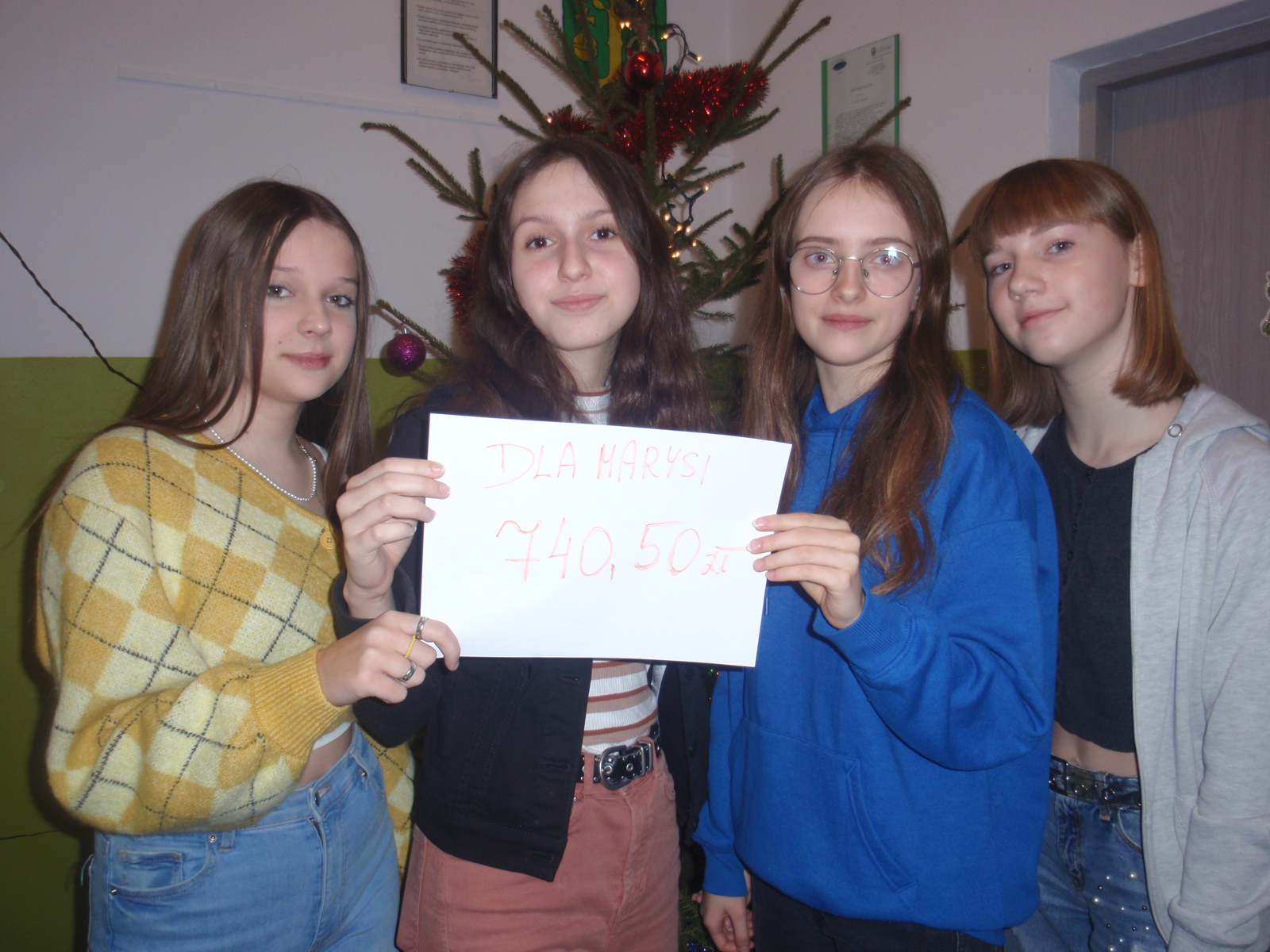 4 dziewczynki  ,trzy  z nich trzymają kartkę z napisem ‘’Dla Marysi ‘’ i napisaną kwotą pieniężną uzbieraną podczas akcji – 740,50 zł.Za uczennicami stoi udekorowana choinka