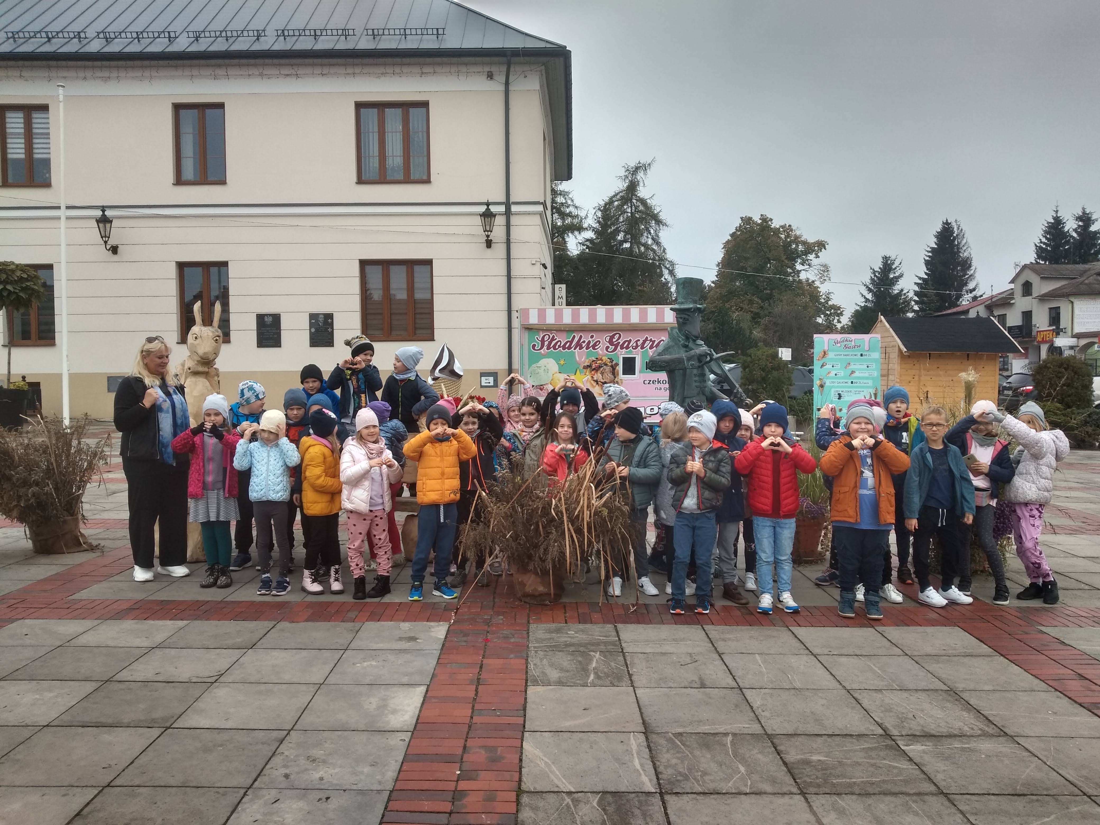 Dzieci ubrane w ciepłe kolorowe kurtki stoją na placu wyłożonym kostką. Za nimi jest wielki biało-beżowy budynek z czarnym dachem, drzewa,drewniany pomnik chrząszcza i małe budki z ciastem. Przed dziećmi stoi bukiet z zasuszonej trawy.